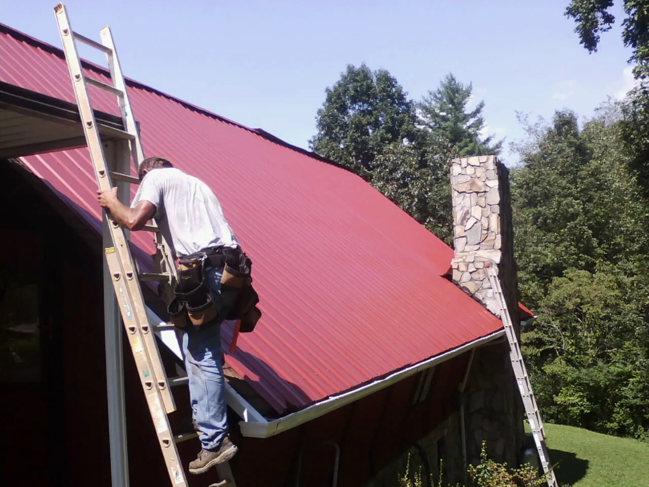 Roofer on a ladder beside a red metal roof and stone chimney — Soffit & Fascia Repair by Roofing Brothers in Mayo
